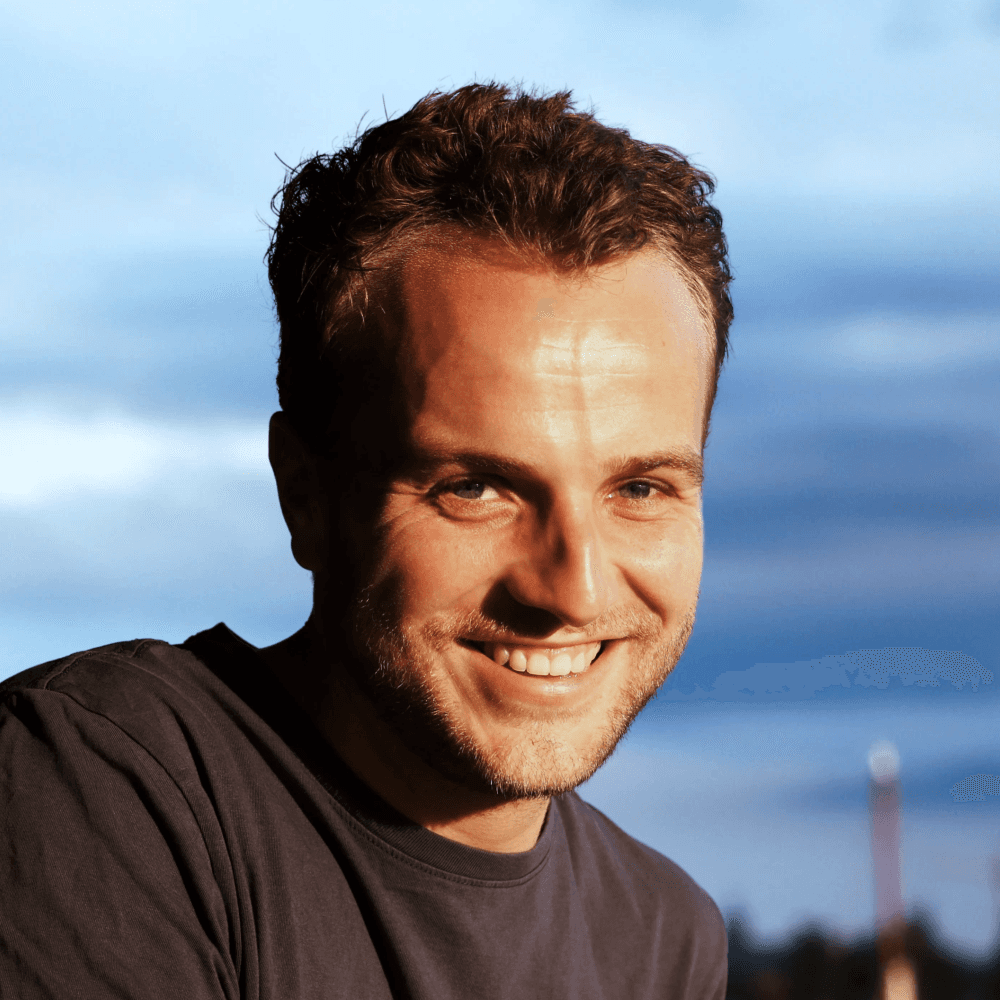 A smiling person with short hair, wearing a dark shirt, against a backdrop of blue sky and clouds.